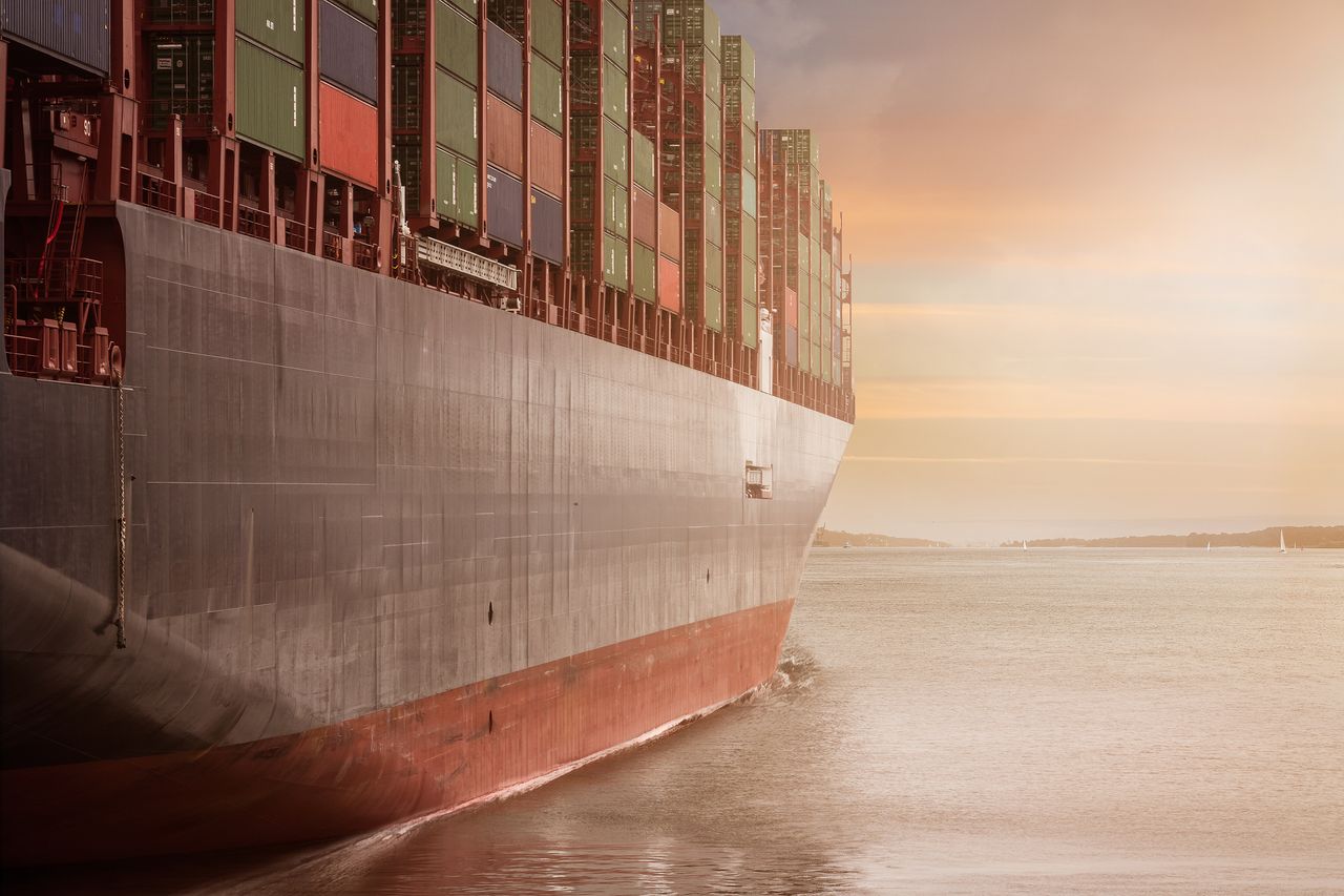 Logistique, Transport & Supply Chain A colossal cargo ship loaded with containers navigates through calm waters against a vibrant sunset sky.